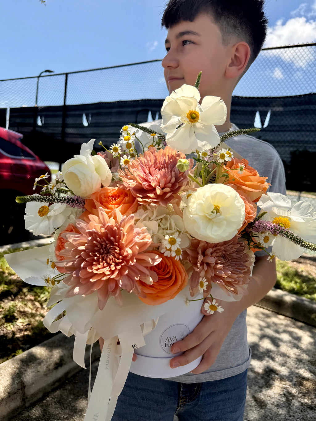 A young boy holding a large, vibrant flower bouquet outdoors. The bouquet features a mix of white, peach, orange, and pink flowers, including dahlia, roses, and ranunculus, wrapped in white paper with a ribbon.
