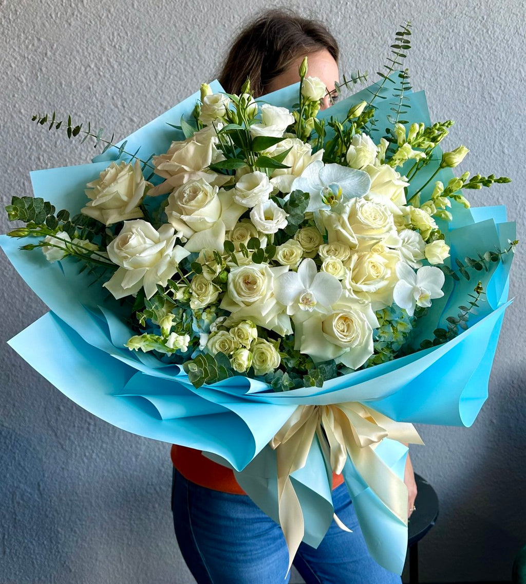 Blue Cloud bouquet with elegant white roses, spray roses, orchids, freesia, and eucalyptus, artistically wrapped in blue paper with satin ribbon — a luxurious floral arrangement full of freshness and sophistication