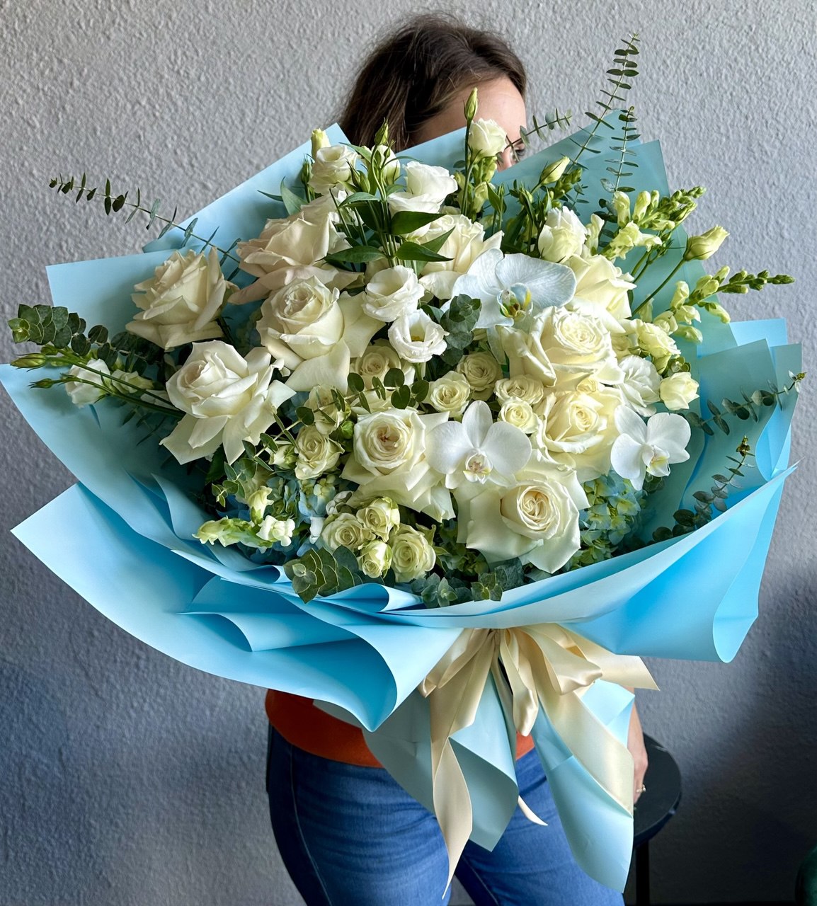 Blue Cloud bouquet with elegant white roses, spray roses, orchids, freesia, and eucalyptus, artistically wrapped in blue paper with satin ribbon — a luxurious floral arrangement full of freshness and sophistication