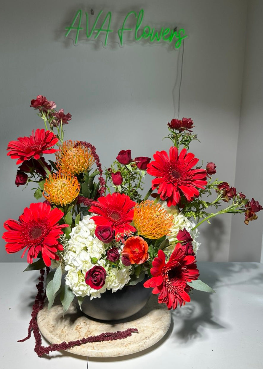 Scarlet Charm flower composition with vibrant red gerberas, exotic orange pincushion protea, white hydrangea, spray roses, and seasonal blooms in a stylish vase — a bold and elegant floral arrangement.